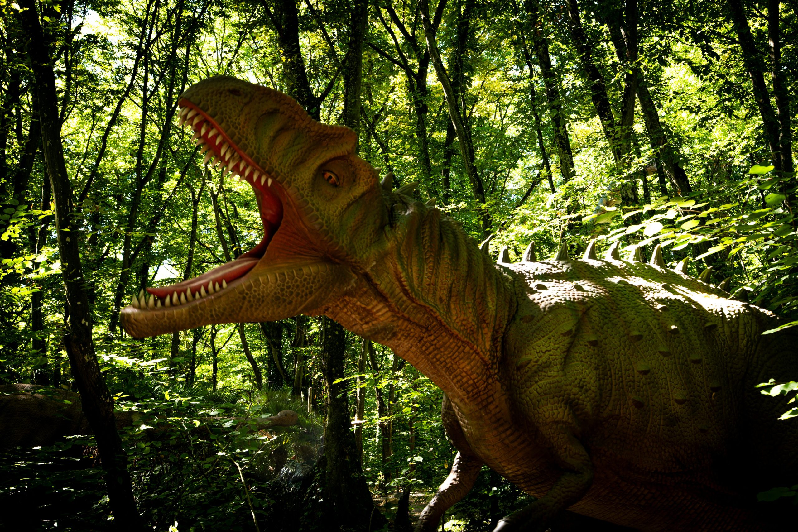 A lifelike dinosaur model roars in a vibrant, sunlit forest setting.